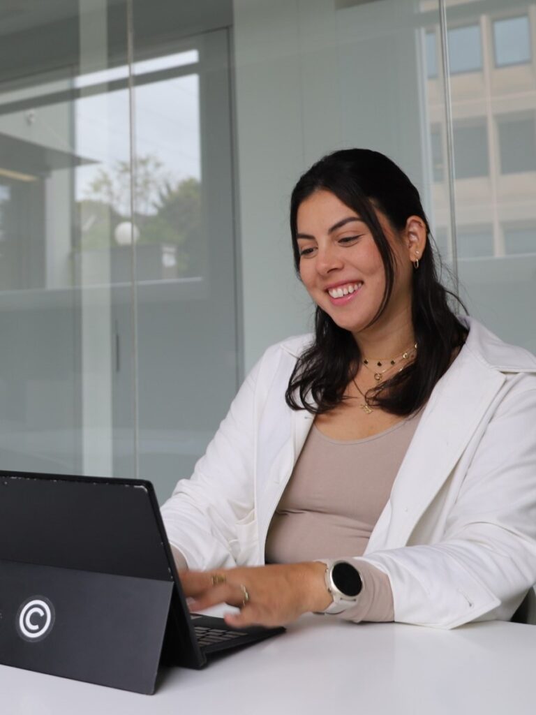 Woman sitting in a modern office at a laptop, smiling and working with focus. She is wearing a white outfit and a smartwatch, with glass walls in the background. 